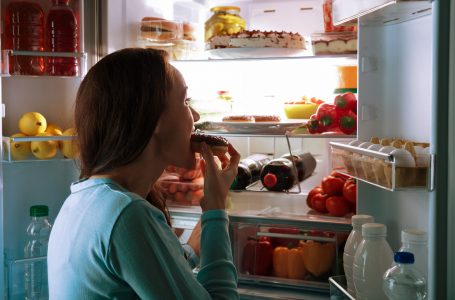 Side View Of A Young Woman Standing In Front Of Refrigerator Eating Donut